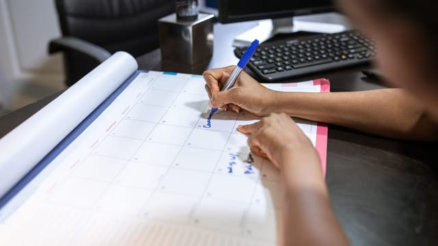 Main qui annote un calendrier mensuel posé sur un bureau